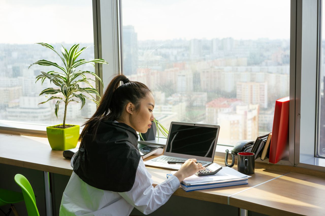 Mastering the First Impression: Your intriguing post title goes here Asian woman focusing on work in a modern high-rise office setting with city view.