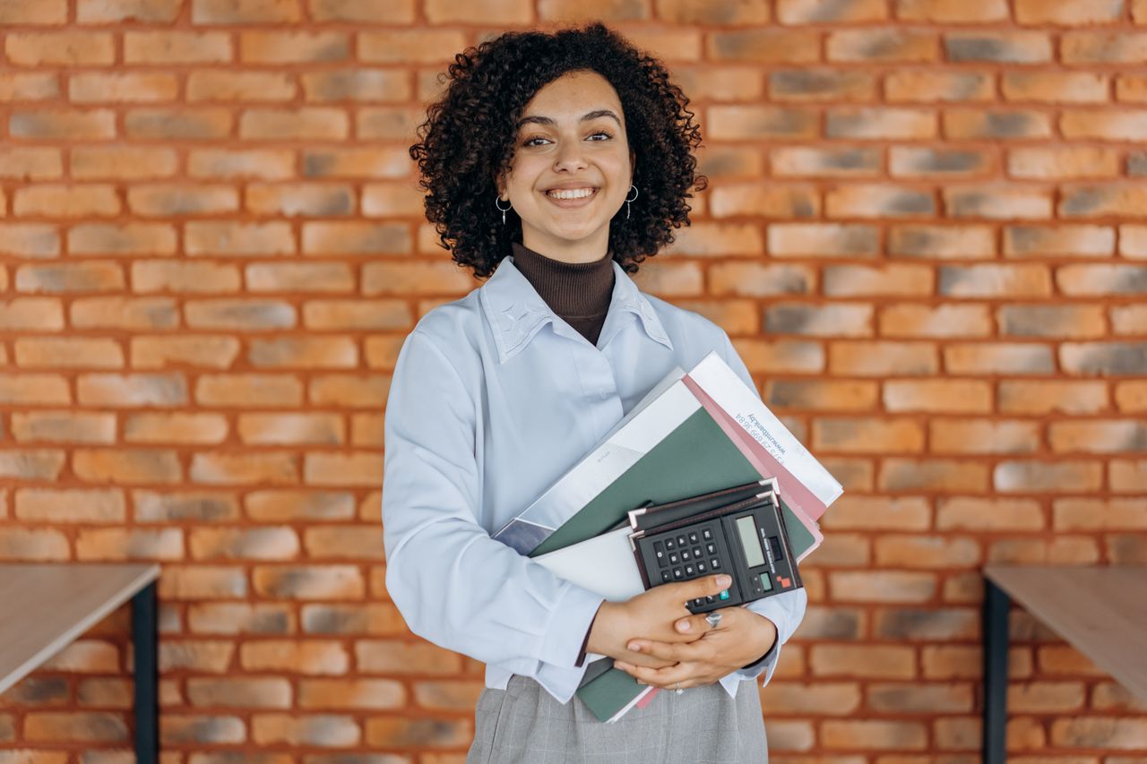 Cheerful woman with curly hair holding folders and a calculator against a brick wall.